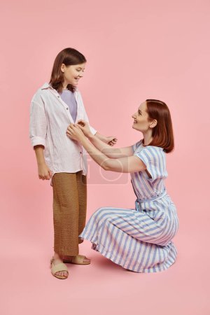 Photo for Mother sitting on haunches and buttoning shirt of preteen girl on pink backdrop, love and care - Royalty Free Image