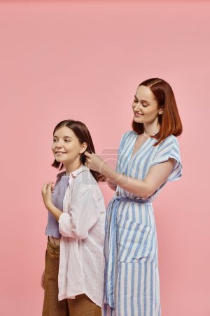 smiling mother braiding hair of happy teenage daughter on pink backdrop in studio, care and love