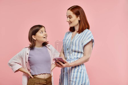 joyful and stylish mother and teenage daughter looking at each other near smartphone on pink