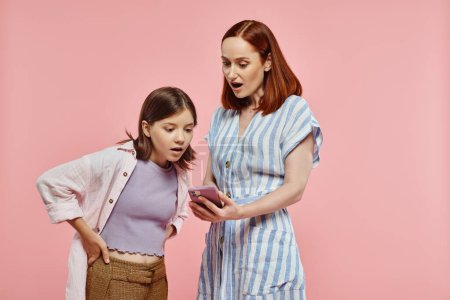 amazed mother and teenage daughter looking at mobile phone while standing on pink backdrop