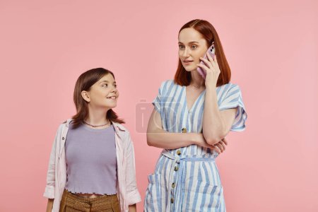 trendy woman talking on mobile phone near smiling preteen girl on pink backdrop, modern mom and teen
