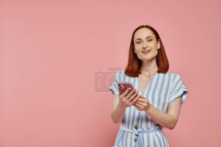 cheerful stylish woman in striped dress holding smartphone and looking at camera on pink backdrop