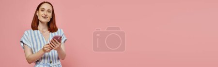 Foto de Mujer alegre y elegante en vestido a rayas con teléfono inteligente y mirando a la cámara en rosa, pancarta - Imagen libre de derechos