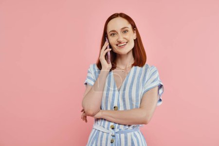 happy and trendy woman in striped dress talking on smartphone and looking at camera on pink