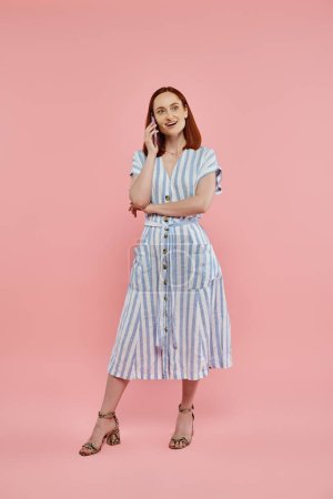 full length of happy and trendy woman in striped dress talking on smartphone on pink backdrop