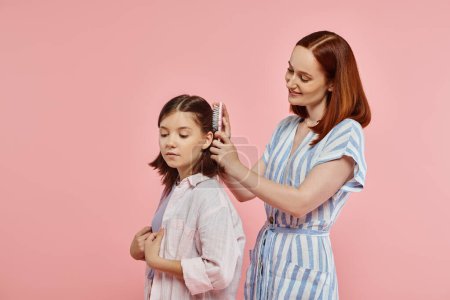 Téléchargez les photos : Mère souriante brossant les cheveux de fille adolescente heureuse sur fond rose en studio, soins et amour - en image libre de droit