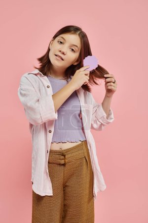 Photo for Preteen girl in stylish clothes brushing hair and looking at camera on pink backdrop, beauty routine - Royalty Free Image