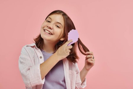 cheerful preteen girl in stylish clothes brushing hair and looking at camera on pink backdrop