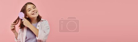 Photo for Happy preteen girl in stylish clothes brushing hair and looking at camera on pink backdrop, banner - Royalty Free Image