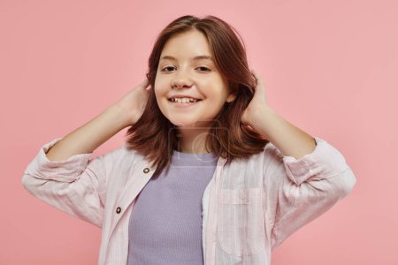 Foto de Alegre y elegante chica preadolescente con el pelo brillante saludable sonriendo a la cámara en el fondo rosa en el estudio - Imagen libre de derechos