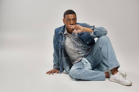 gut aussehender junger afrikanisch-amerikanischer Mann im trendigen Jeans-Outfit auf dem Boden sitzend, Modekonzept