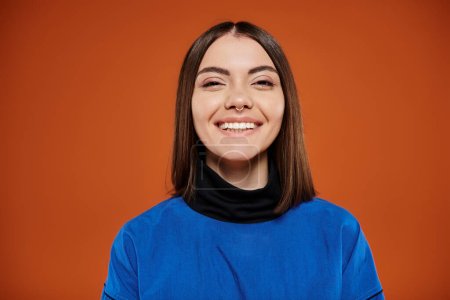 happy young woman with pierced nose looking at camera and smiling on orange backdrop, blue jacket