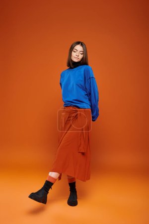 fall season attire, attractive woman in skirt and blue sweatshirt standing on orange backdrop