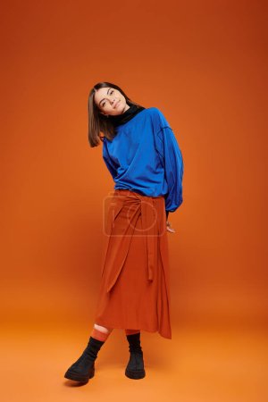 Photo for Fall season attire, beautiful woman in skirt and blue sweatshirt standing on orange backdrop - Royalty Free Image