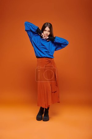 autumn fashion, attractive woman in skirt, blue sweatshirt and boots standing on orange backdrop