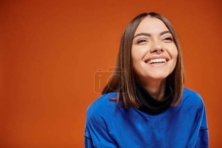 glückliche junge Frau mit durchbohrter Nase, die wegschaut und auf orangefarbenem Hintergrund lächelt, blaues Sweatshirt