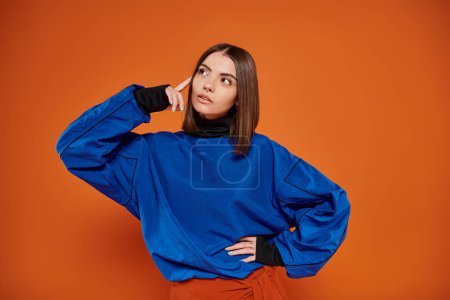 nachdenkliche junge Frau mit durchbohrter Nase, die mit der Hand an der Hüfte auf orangefarbenem Hintergrund steht, schaut weg