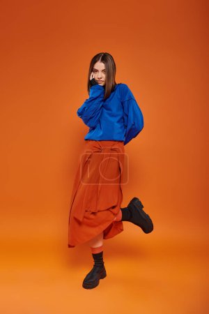 Foto de Mujer de moda y joven con la nariz perforada de pie en la falda de otoño y botas en el fondo naranja - Imagen libre de derechos