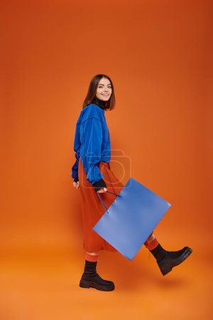 glückliche Frau in herbstlicher Kleidung, die mit Einkaufstasche auf orangefarbenem Hintergrund spaziert, Black-Friday-Konzept