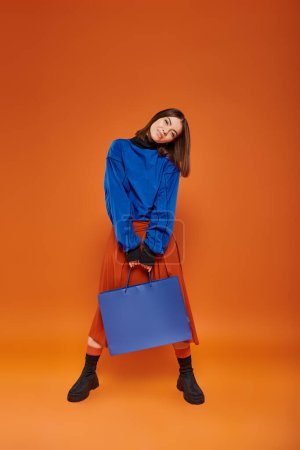 brünette Frau in Herbstkleidung mit Einkaufstasche vor orangefarbenem Hintergrund, Black-Friday-Konzept