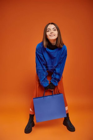 cheerful woman in autumn attire standing with shopping bag on orange backdrop, black friday concept