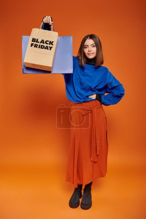 cheerful woman in autumn attire standing with shopping bags on orange backdrop, black friday concept