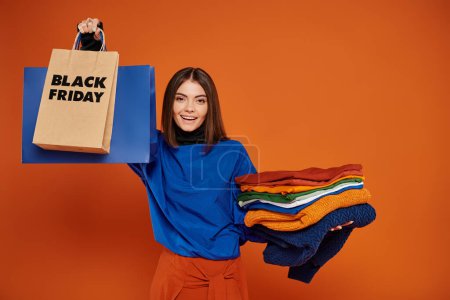 joyful woman holding shopping bags and stack of clothes on orange backdrop, black friday concept