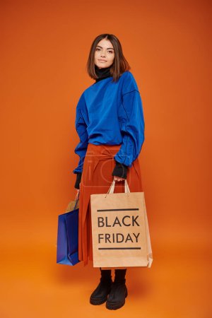 junge fröhliche Frau im herbstlichen Outfit mit Einkaufstüten vor orangefarbenem Hintergrund, Black Friday Sale