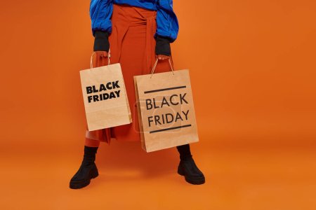 gestutzte Frau in Herbstrock und Stiefeln mit Einkaufstüten vor orangefarbenem Hintergrund, schwarzer Freitag