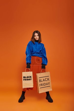 Foto de Longitud completa, mujer morena en traje de otoño sosteniendo bolsas de compras sobre fondo naranja, viernes negro - Imagen libre de derechos