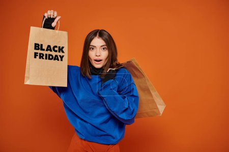 Schockierte Frau mit Einkaufstüten mit schwarzen Freitagsbriefen auf orangefarbenem Hintergrund, Verkaufssaison