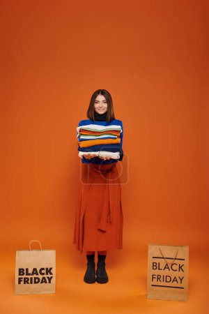 mujer alegre sosteniendo pila de ropa de abrigo cerca de bolsas de compras con letras de viernes negro en naranja