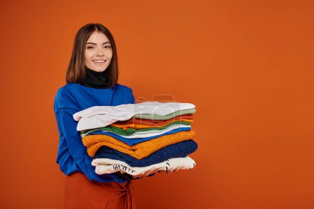 cheerful woman holding stack of warm clothes and smiling on orange backdrop, black friday concept