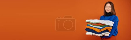 Téléchargez les photos : Femme gaie tenant pile de vêtements chauds et souriant sur fond orange, bannière vendredi noir - en image libre de droit