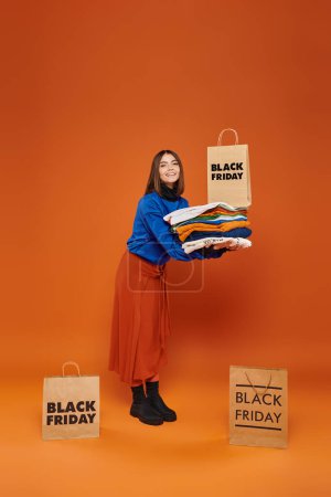 Foto de Mujer positiva sosteniendo pila de ropa de abrigo y bolso de compras en fondo naranja, viernes negro - Imagen libre de derechos