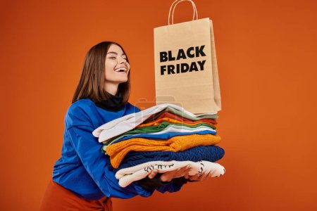mujer alegre sosteniendo pila de ropa de abrigo y bolso de compras en fondo naranja, viernes negro