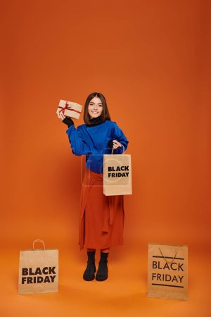 joyful woman holding wrapped present and shopping bag on orange backdrop, black friday sales