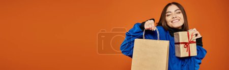 pleased woman holding wrapped present and shopping bag on orange backdrop, black friday banner