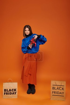 Téléchargez les photos : Femme heureuse tenant présent enveloppé près des sacs à provisions sur fond orange, noir vendredi ventes - en image libre de droit