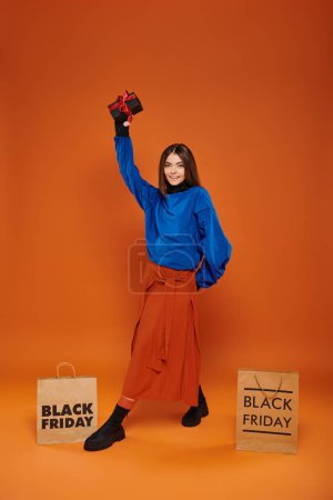 cheerful woman holding wrapped present near shopping bags on orange backdrop, black friday discounts
