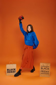 cheerful woman holding wrapped present near shopping bags on orange backdrop, black friday discounts Poster #680598378