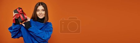 Photo for Cheerful woman with brunette hair holding wrapped present on orange backdrop, black friday banner - Royalty Free Image