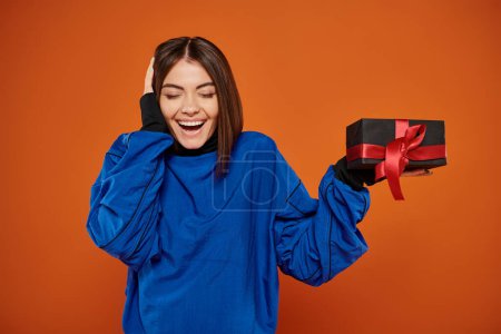 glückliche Frau mit brünetten Haaren hält eingewickeltes Geschenk auf orangefarbenem Hintergrund, schwarzer Freitag