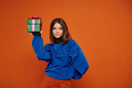 schöne Frau mit brünetten Haaren hält eingewickeltes Geschenk auf orangefarbenem Hintergrund, Frohe Weihnachten