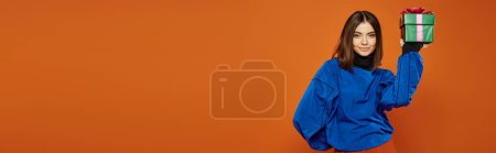young woman with brunette hair holding wrapped present on orange background, Merry Christmas banner