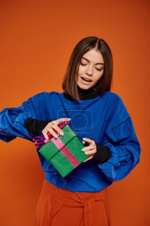 hübsche Frau mit brünetten Haaren Öffnung eingewickelt Geschenk auf orangefarbenem Hintergrund, Frohe Weihnachten