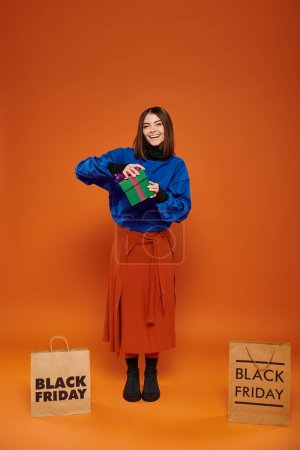 glückliche Frau öffnet Geschenk in der Nähe von Einkaufstaschen mit schwarzen Freitagsbuchstaben auf orangefarbenem Hintergrund