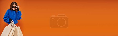 Photo for Woman wearing stylish sunglasses and holding shopping bags on orange backdrop, black friday banner - Royalty Free Image