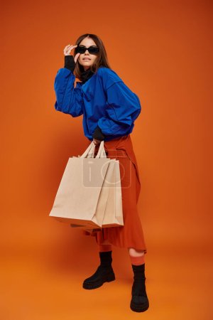 Foto de Morena mujer con gafas de sol con estilo y la celebración de bolsas de compras sobre fondo naranja, viernes negro - Imagen libre de derechos