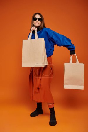 brünette Frau mit stylischer Sonnenbrille und Einkaufstüten vor orangefarbenem Hintergrund, Black Friday Sale